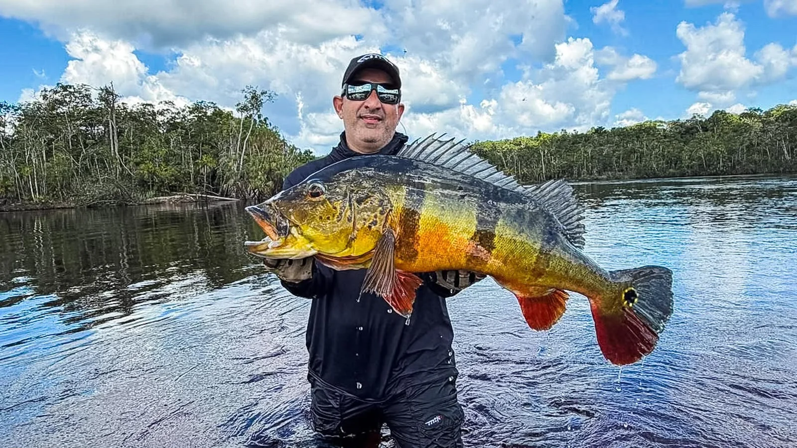 Pescador segura um grande tucunaré, com a água e a floresta ao fundo